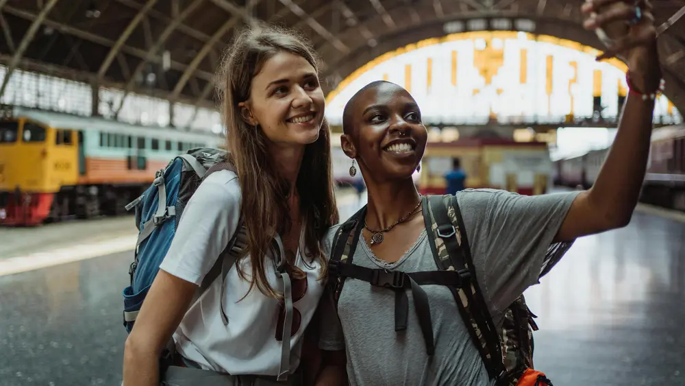 Two young women take a selfie with a smartphone inside a European train terminal.