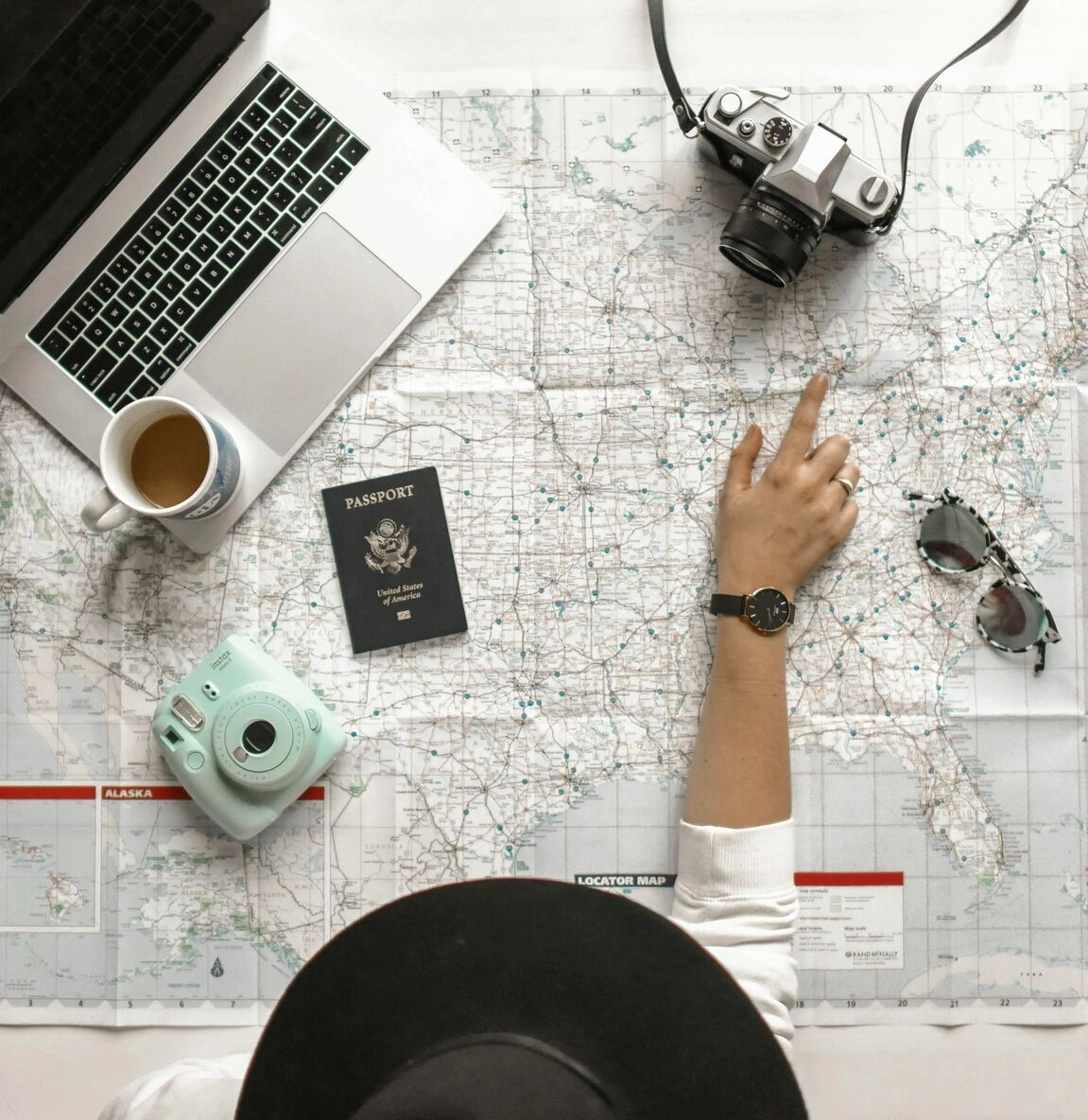 hand pointing to a world map with a passport, polaroid camera, sunglasses, coffee and a laptop computer